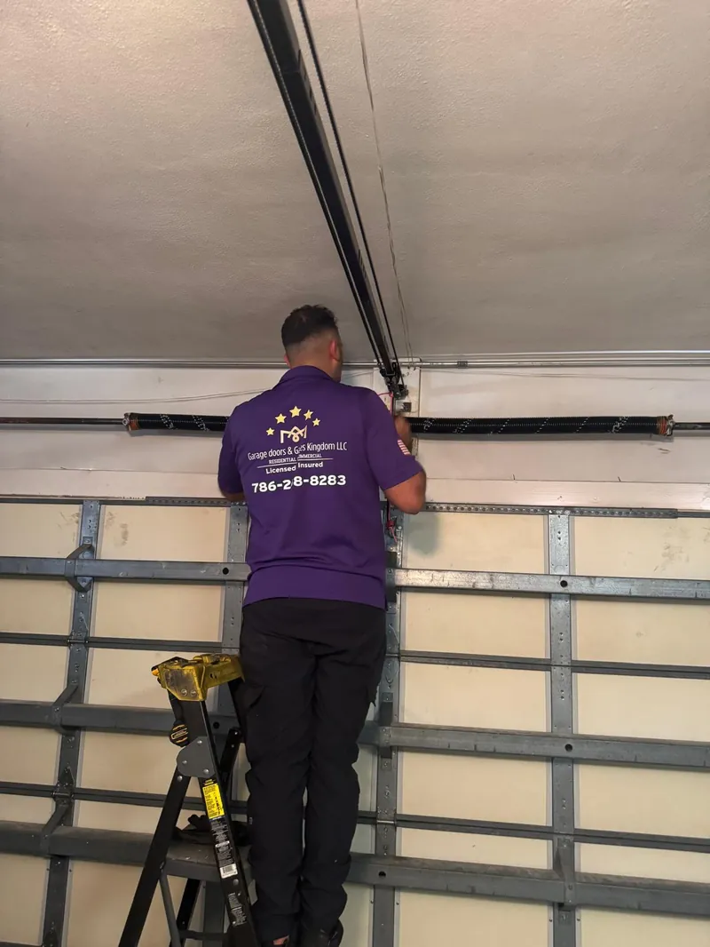 Back view of Garage Door Kingdom licensed and insured technician in branded purple uniform displaying the 786-258-8283 phone number while working on a garage door